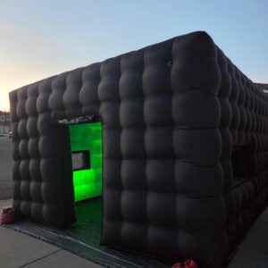 Black inflatable cube structure with green-lit interior at dusk.